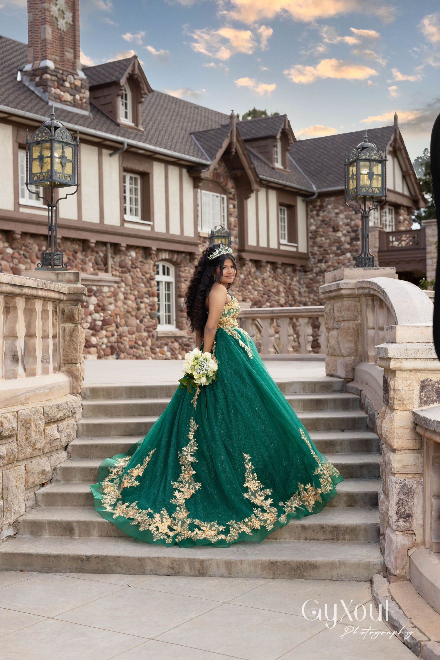 An emerald green ball gown with gold embellishments sparkles on stone steps at a luxurious estate during golden hour.