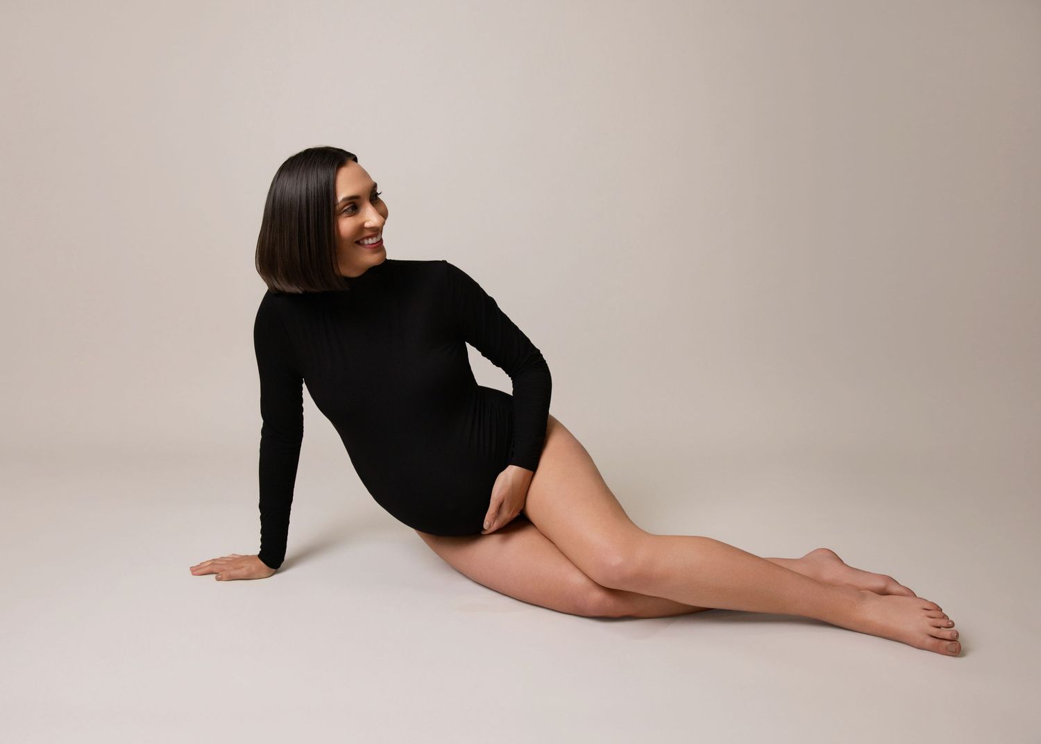 Woman in black long sleeve bodysuit poses elegantly on beige studio floor during maternity photo session.