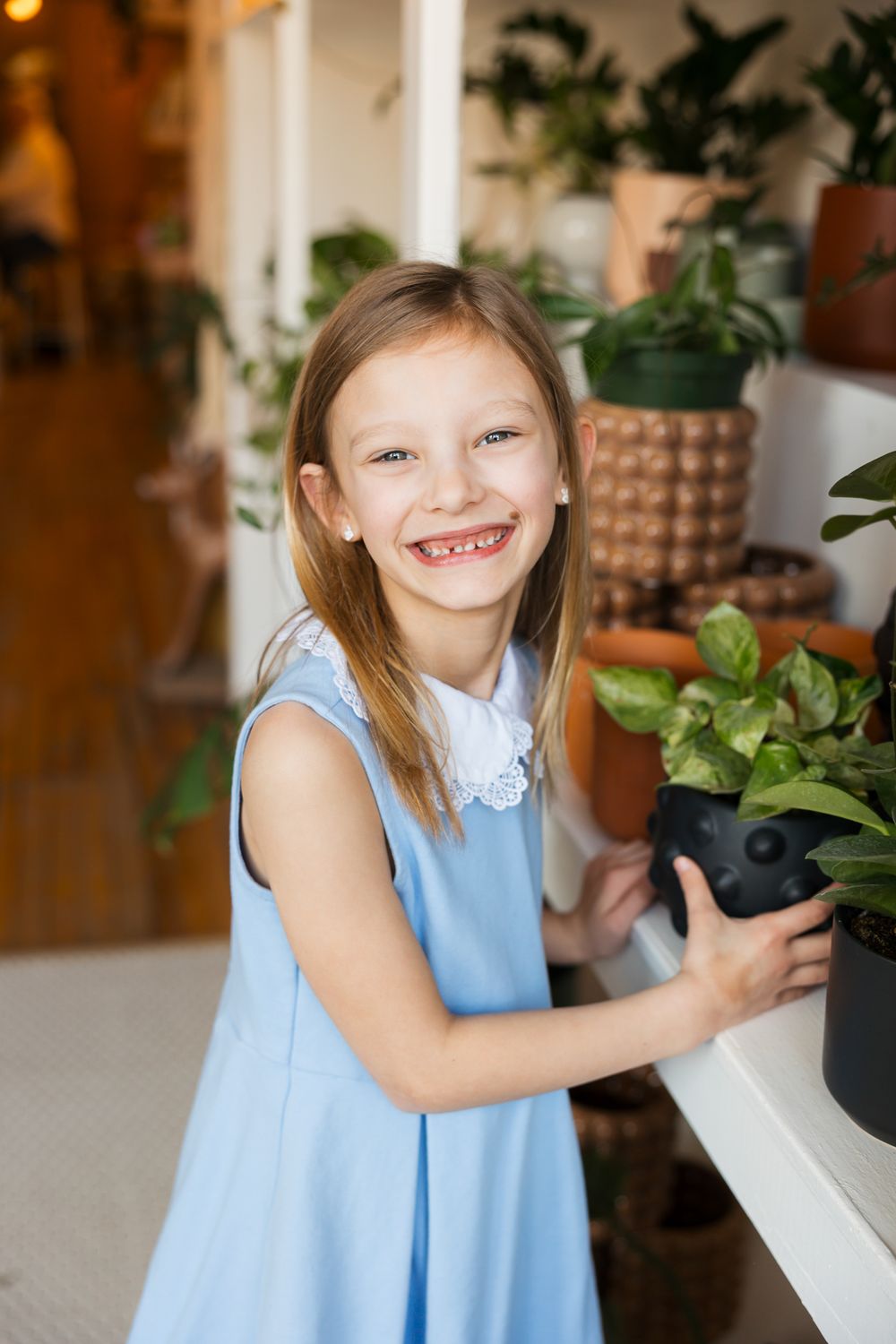 A young person in a blue sleeveless dress smiles while holding a potted plant in a room with houseplants and decor.