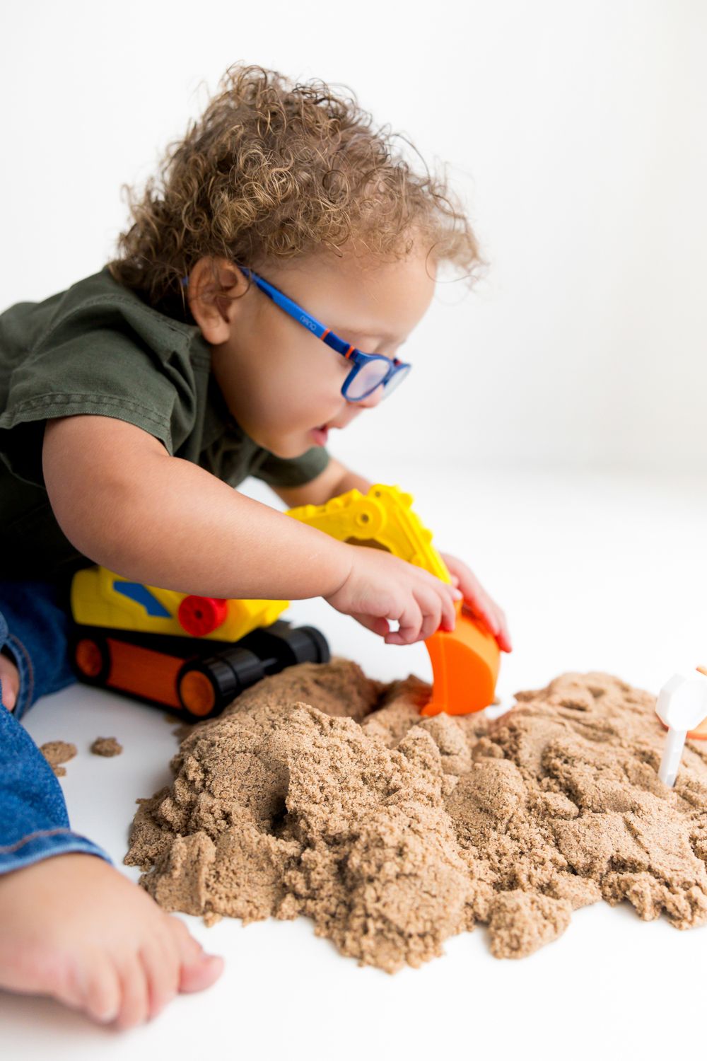 Young child wearing blue glasses plays with kinetic sand and toys on a white surface during indoor playtime.