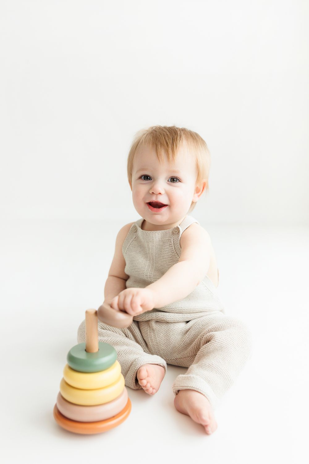Happy toddler in beige romper plays with colorful wooden stacking rings toy on white background.
