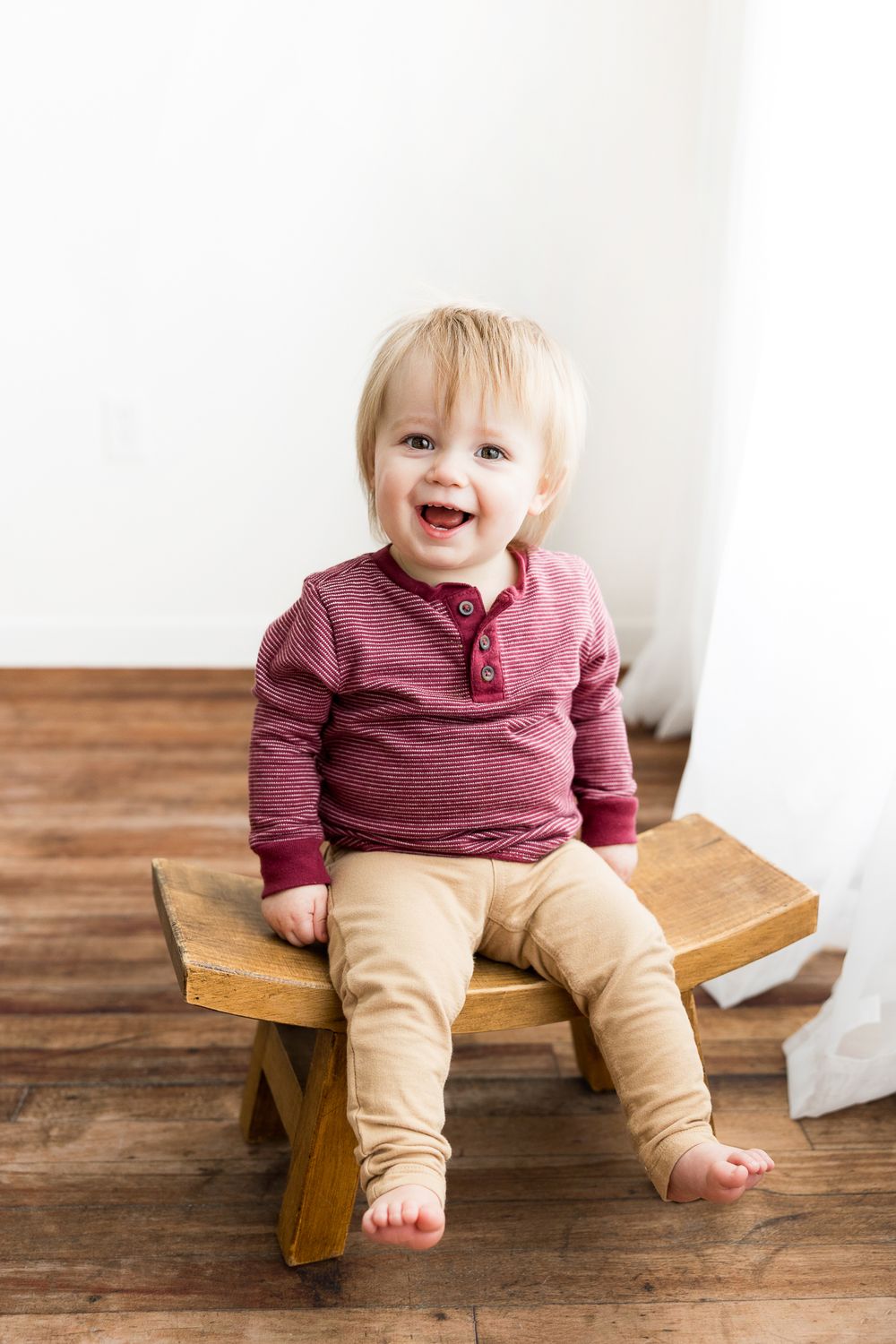 A toddler in a burgundy shirt and khaki pants sits smiling on a wooden bench against a white wall.