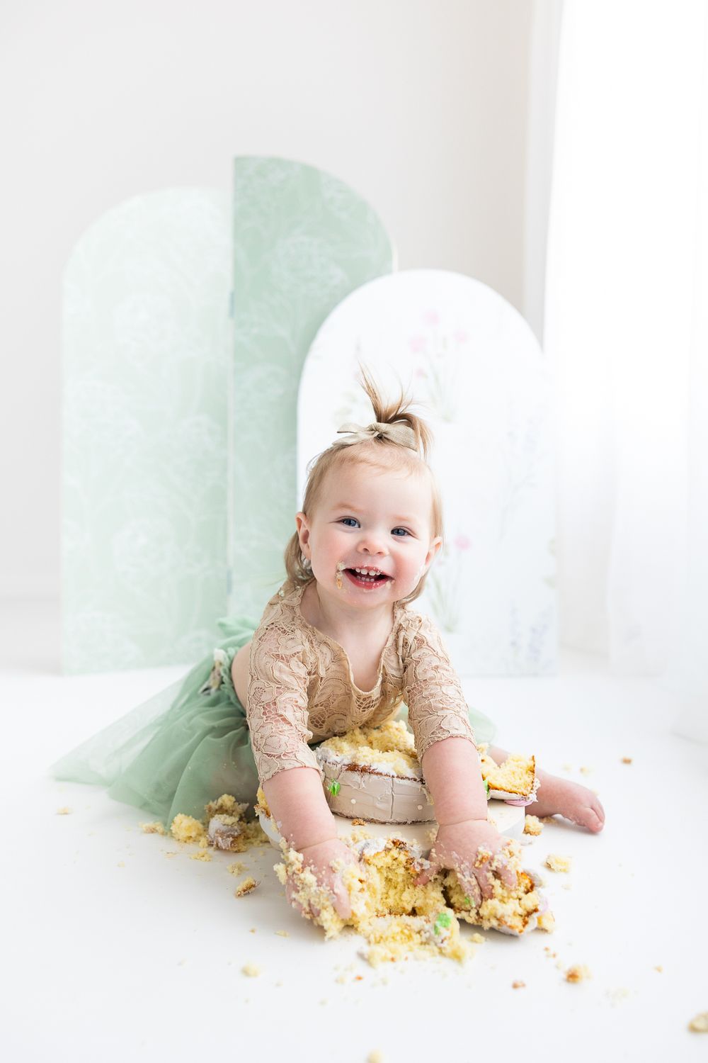 Baby wearing beige lace dress sits on white floor with mint green backdrop playing with smashed yellow cake.