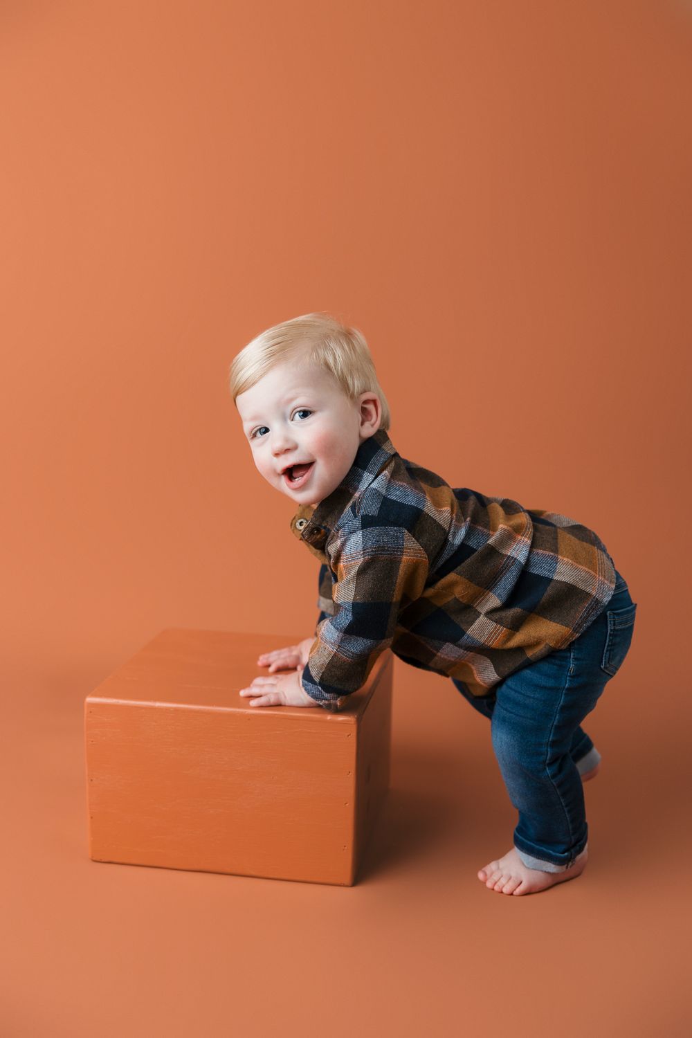 Toddler wearing plaid flannel shirt and jeans plays with orange box against matching orange background.