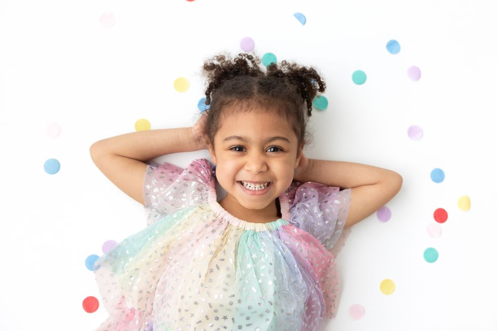 Young person in a sparkly rainbow dress laying on floor with colorful confetti scattered around.
