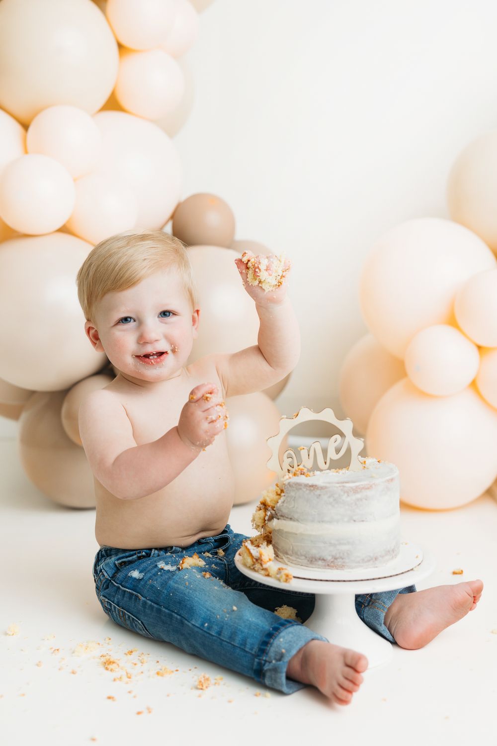 A joyful cake smash photography session with peach balloons and a small grey cake on white backdrop.