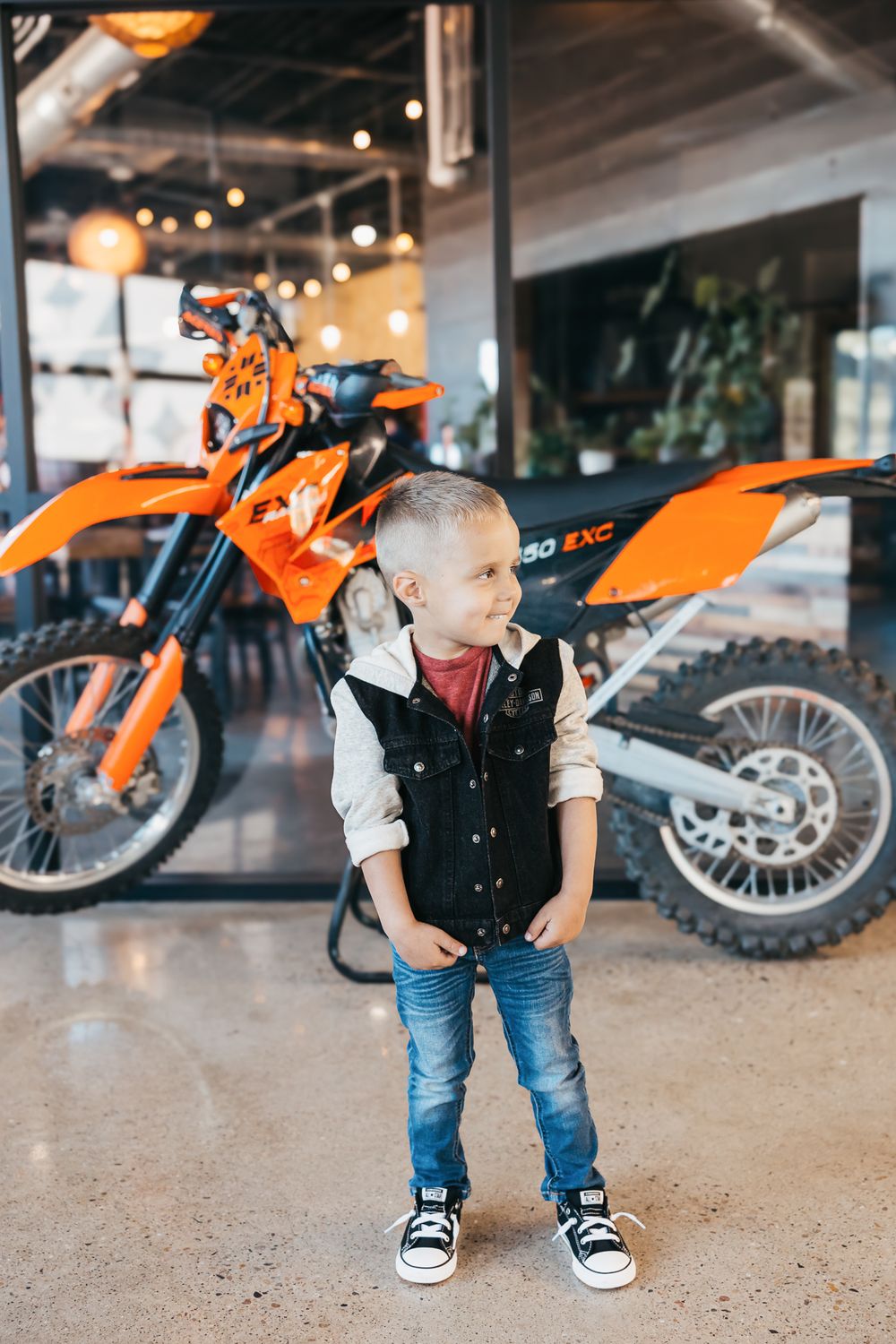 Young child in denim and black vest stands confidently next to bright orange KTM dirt bike in showroom setting.