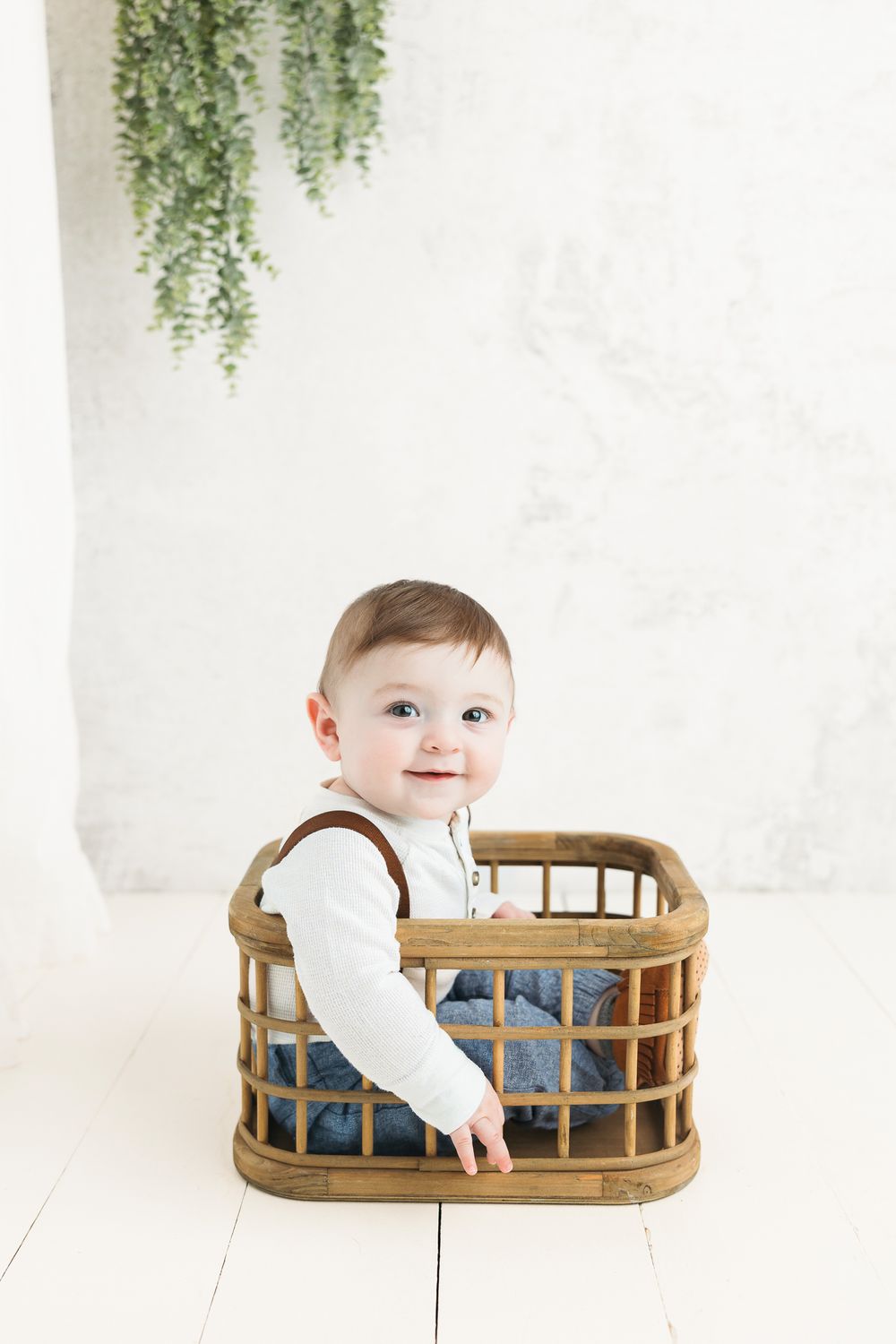 Small toddler sitting in a wooden crate against white backdrop with hanging greenery in minimalist studio setting.