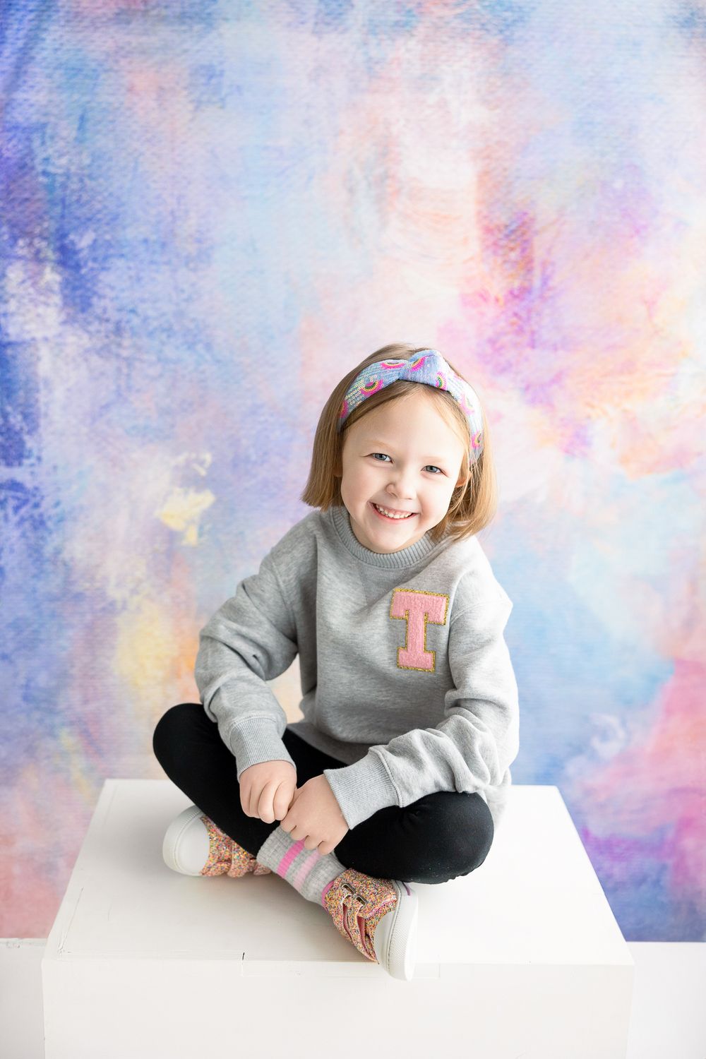 Child seated cross-legged on white platform against dreamy blue and pink watercolor backdrop.