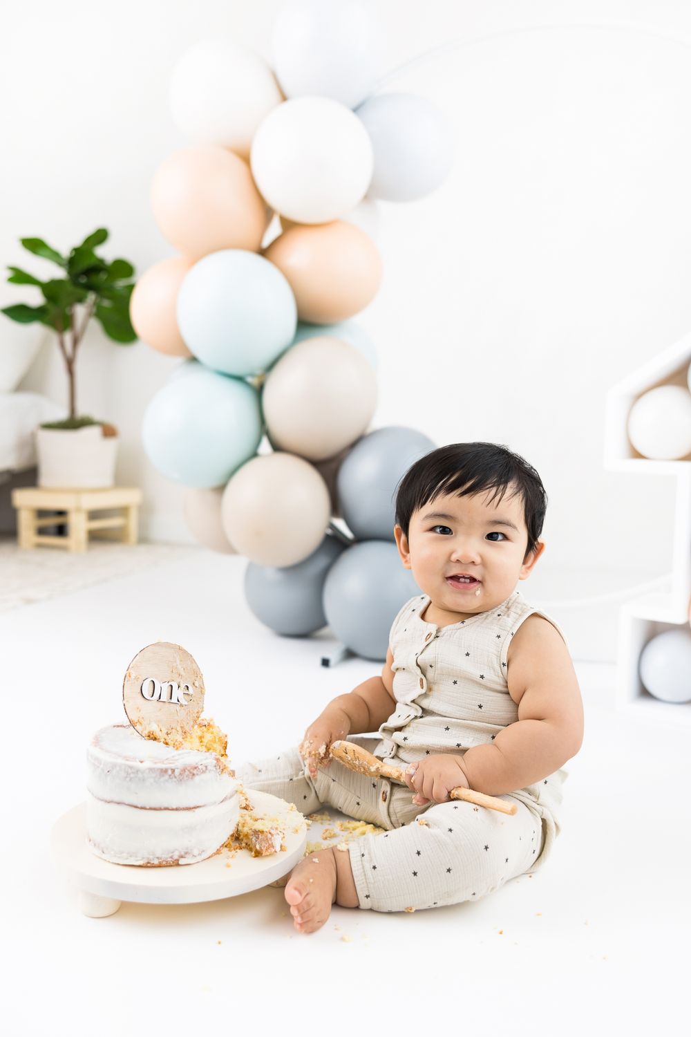 Baby in polka dot outfit sits by pastel balloon arch and cake during first birthday photoshoot.