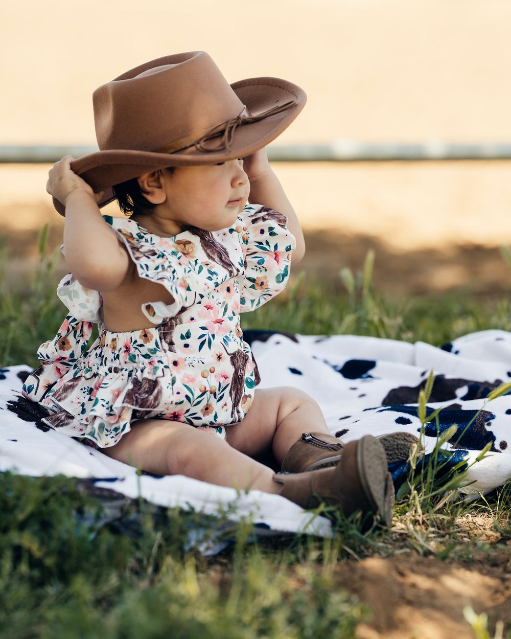 A small figure wearing a floral outfit and brown cowboy hat relaxes on a blanket in the grass.