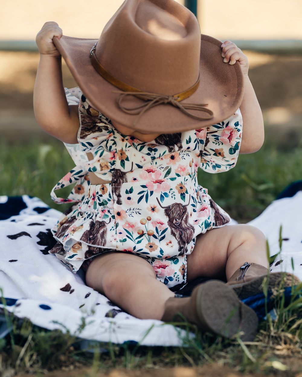 A brown cowboy hat and floral outfit are arranged on grass with a white blanket.