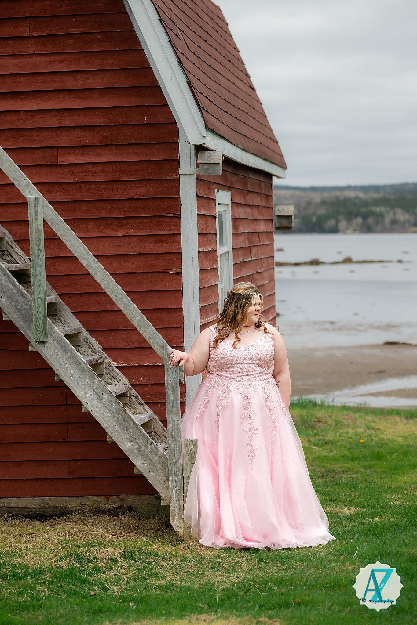 A pink lace gown stands elegantly against a red coastal building with stairs overlooking a scenic beach landscape.