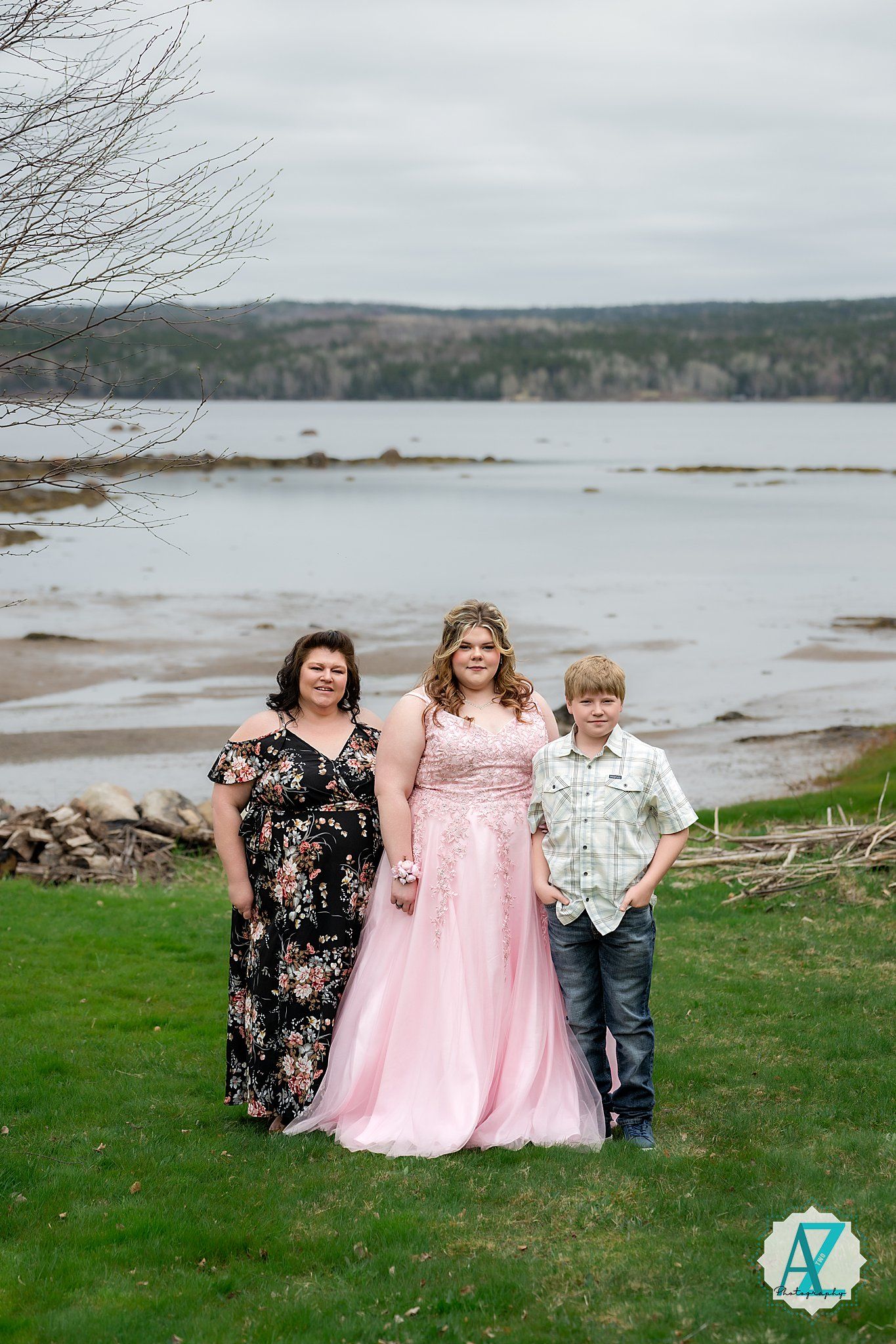 Family group in formal wear poses together on grassy shore with coastal water and hills in background.