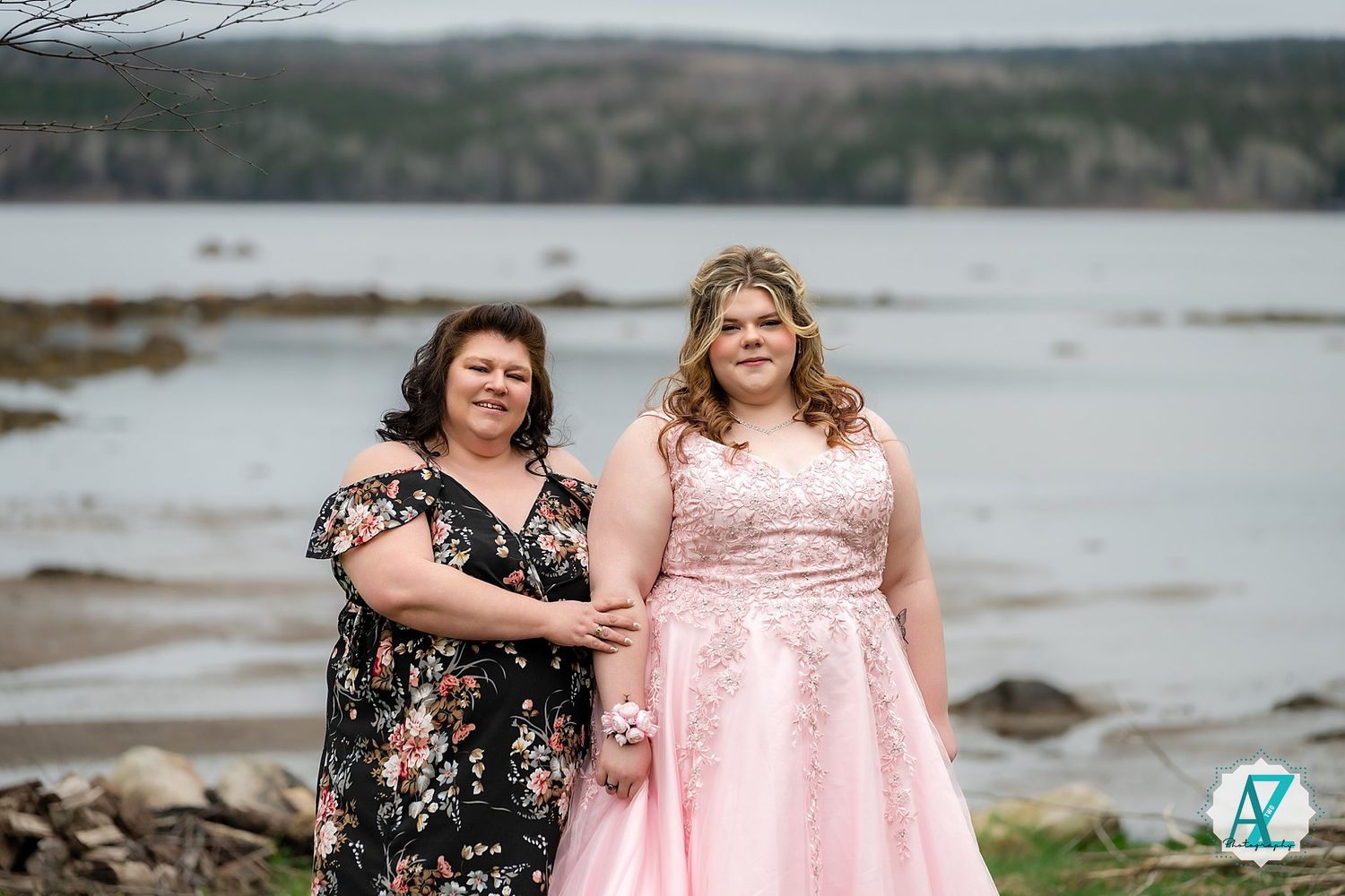 Two people in elegant dresses stand together on a rocky shore with a serene lake and forest backdrop.
