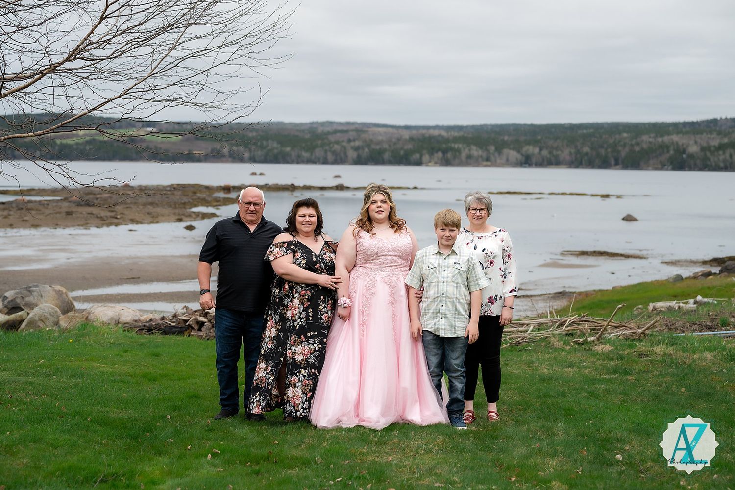 A group stands together on green grass near a scenic lake with hills in background on an overcast day.