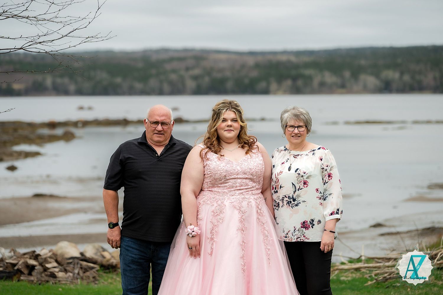 Three people pose for a photo by a lake with someone wearing a pink formal dress in the center.