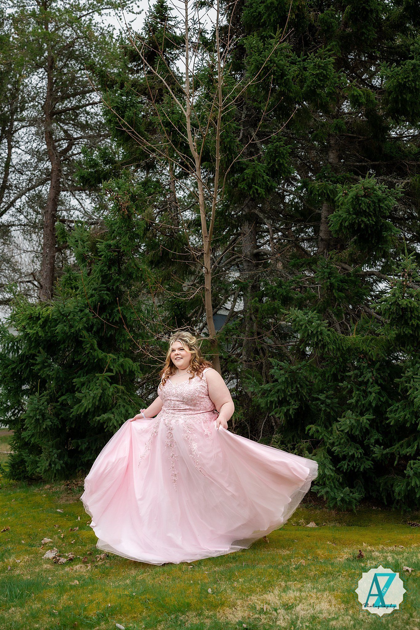 A person in a flowing pink formal gown twirls in a garden setting with evergreen trees in the background.