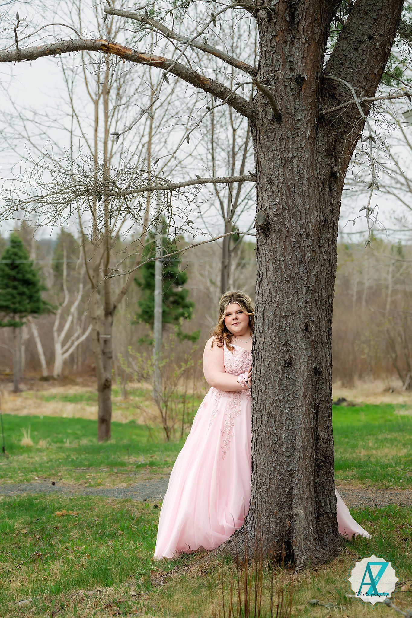 A person in a flowing pink gown stands beside a large tree trunk in a springtime outdoor setting.