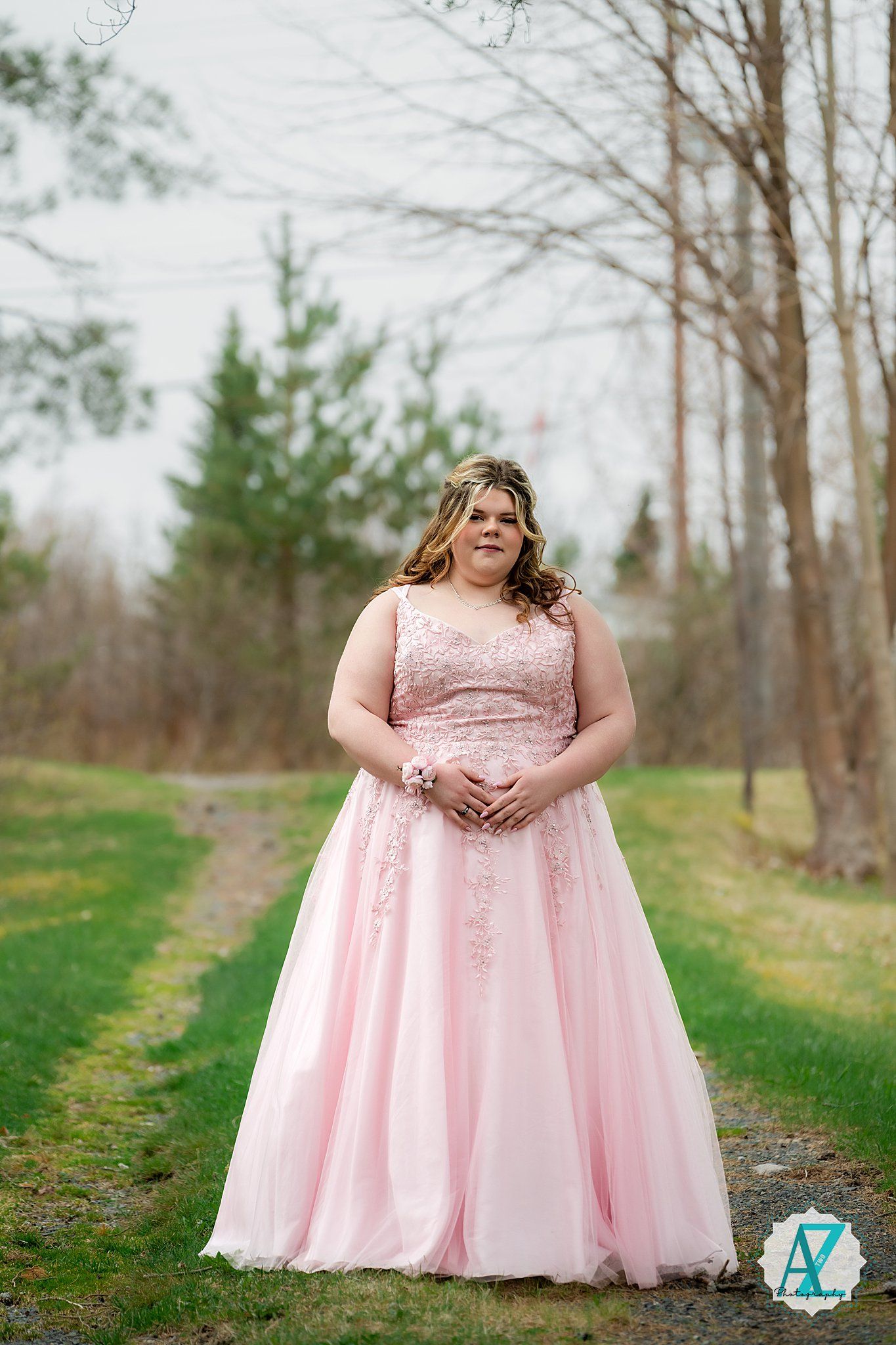A person wearing a blush pink formal gown with lace details stands outdoors on a grassy path among bare trees.