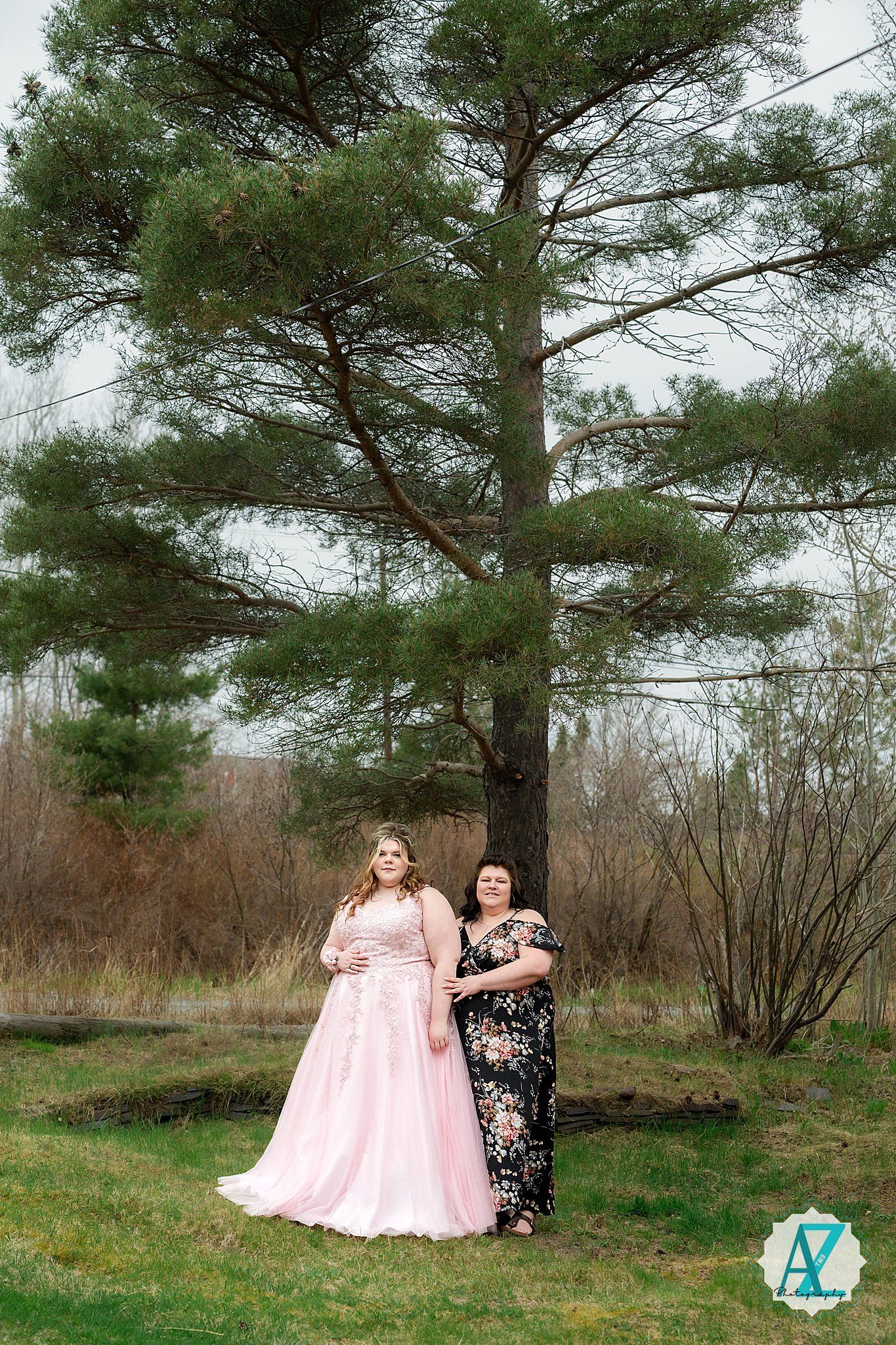 Two people in formal attire pose beneath a towering pine tree in a natural outdoor setting.