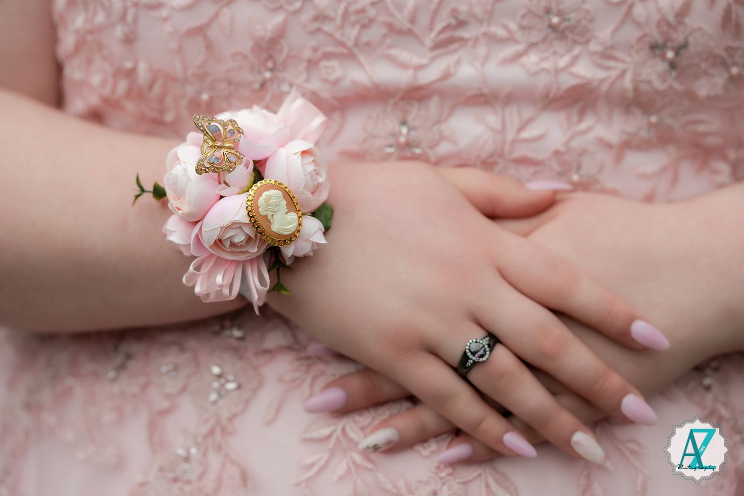 A delicate pink floral corsage adorns a wrist against soft pink fabric, with a silver ring visible on the hand.