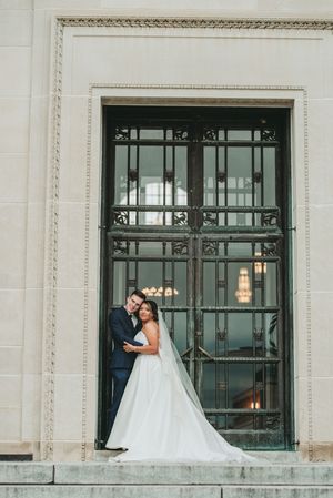 Wedding couple poses elegantly against a stone building with ornate black window frames.