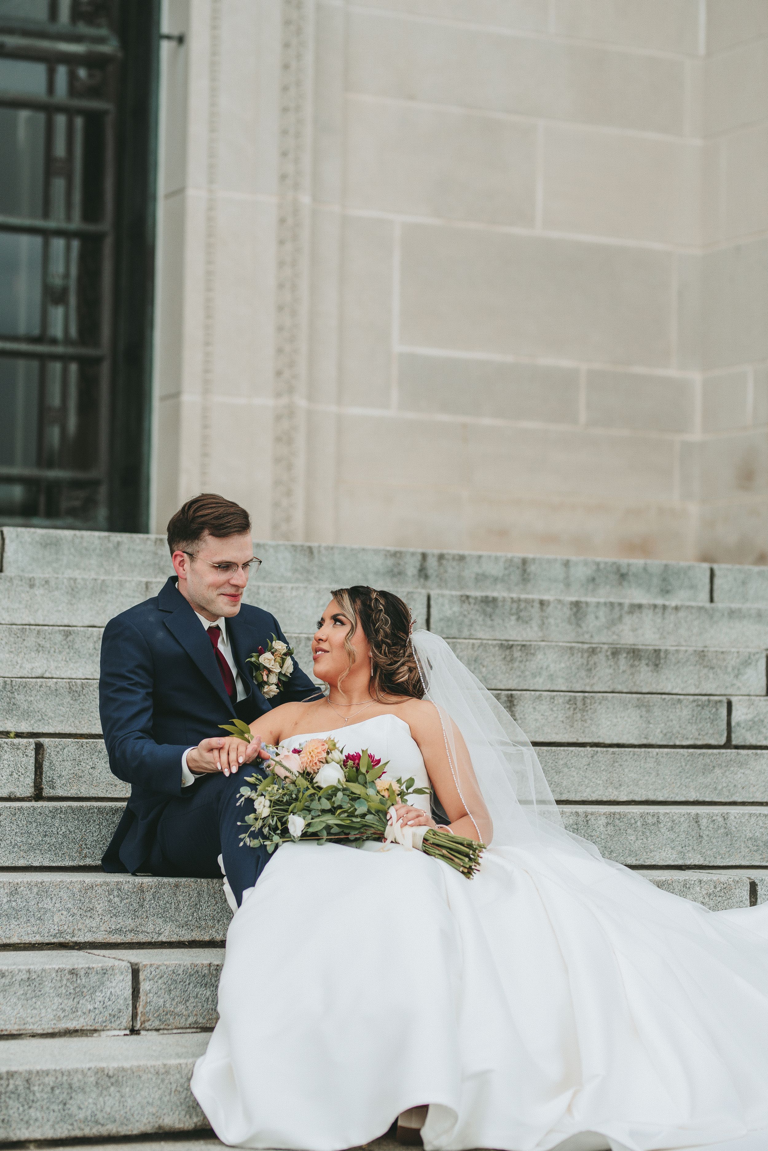 Bride in flowing white gown sits gracefully on stone steps with groom.