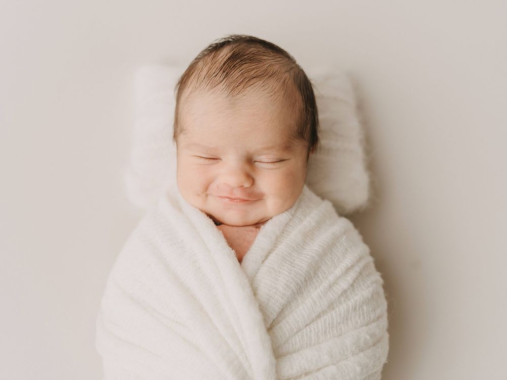 Sleeping newborn snuggled in soft white blanket smiling contentedly during professional photo session.