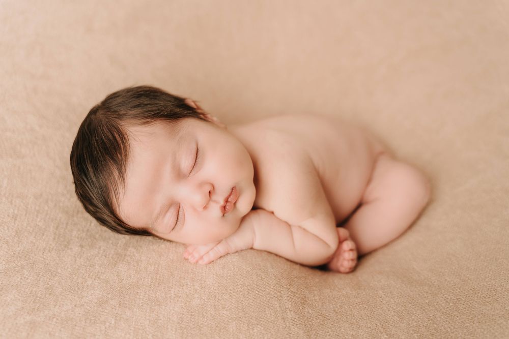 Peaceful sleeping newborn baby curled up on soft beige backdrop during professional photo session.