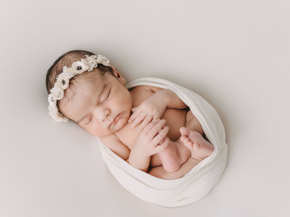 Sleeping baby wrapped in soft white fabric wearing delicate floral headband on neutral background.