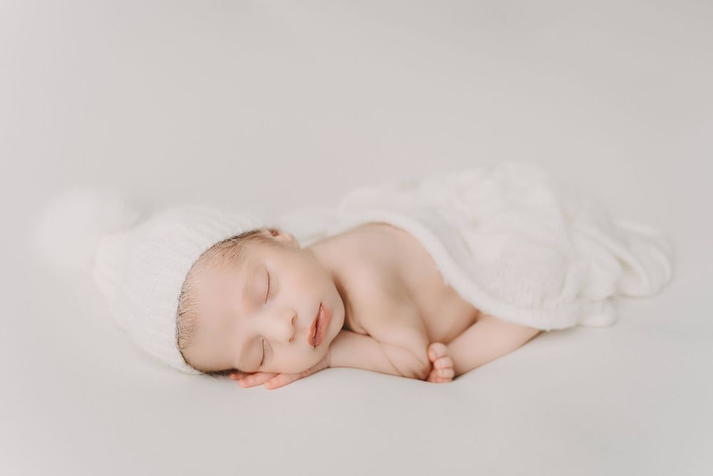 A peaceful sleeping newborn wrapped in soft white fabric lies curled up on a light backdrop, creating a serene portrait.