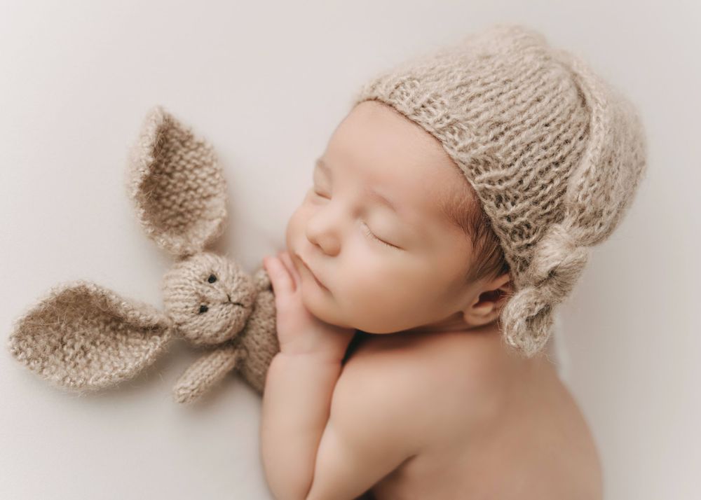 A sleeping newborn wearing a knit beanie lies on a soft surface with a plush bunny toy.