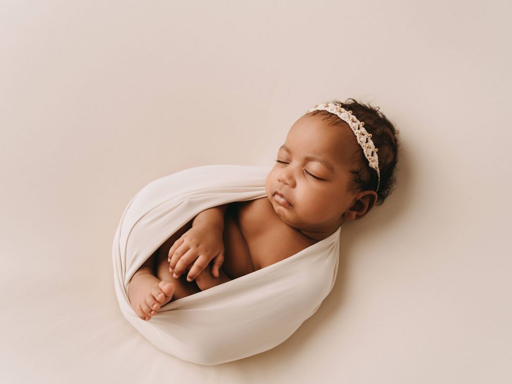 Sleeping newborn baby wrapped in soft white fabric wearing delicate headband rests peacefully in curved pose.