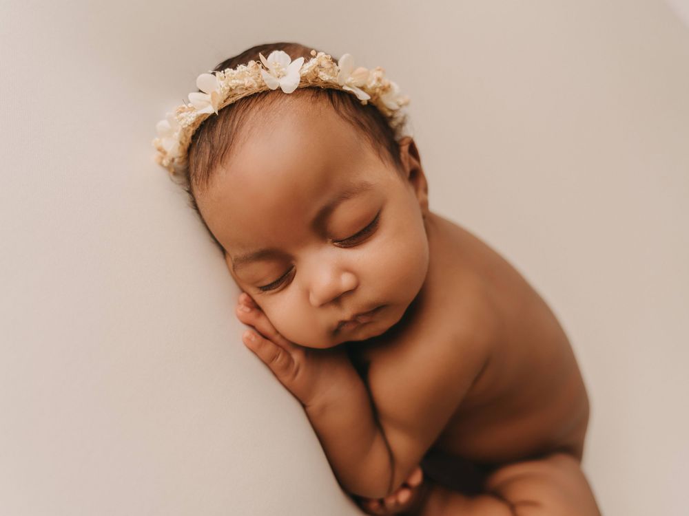 Peaceful sleeping newborn baby wearing floral headband resting on beige background during professional photo session.