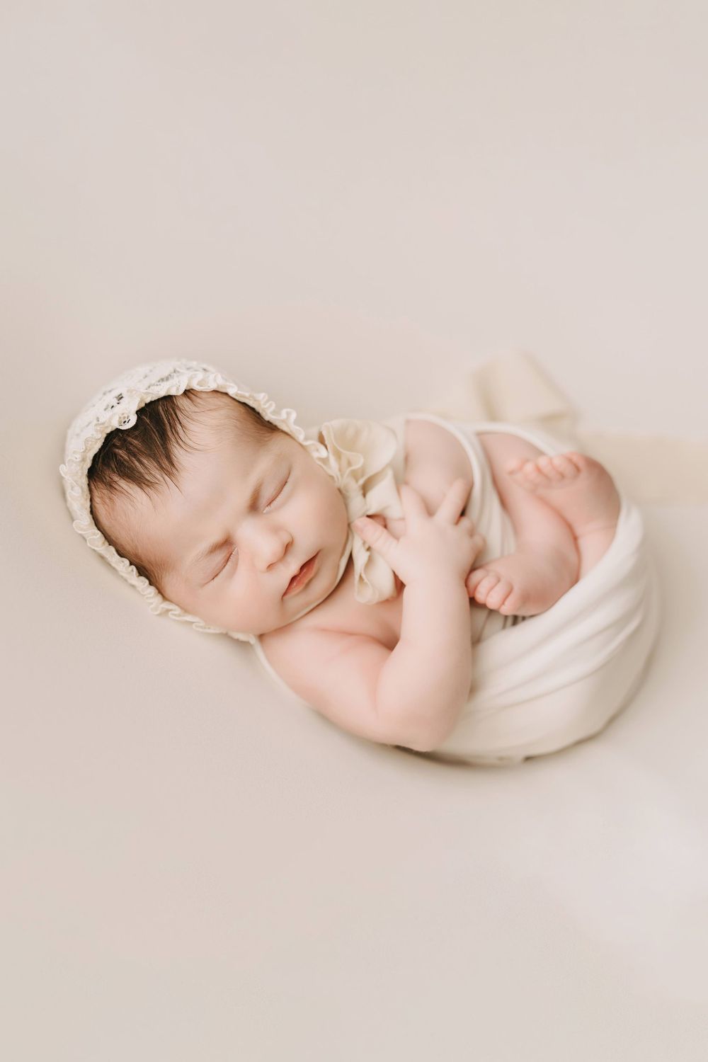 Peaceful sleeping newborn in cream bonnet wrapped in soft white swaddle against neutral backdrop.