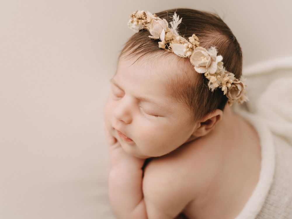Newborn wearing delicate floral crown headband while sleeping on cream backdrop.