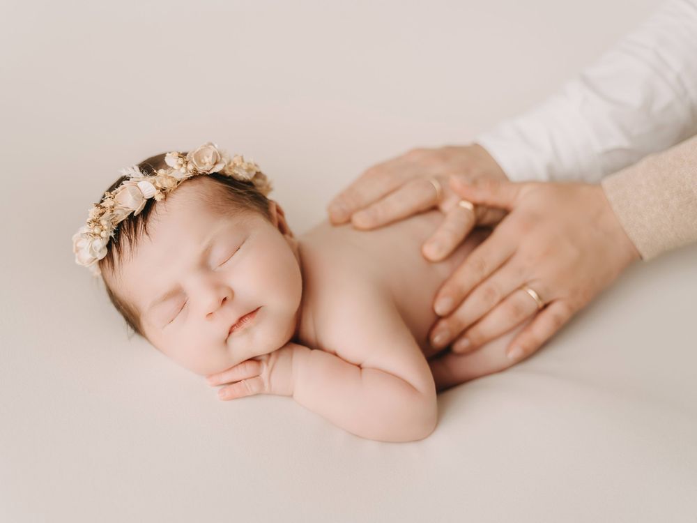 Peaceful sleeping newborn wearing floral crown with gentle adult hand providing comfort on neutral background.