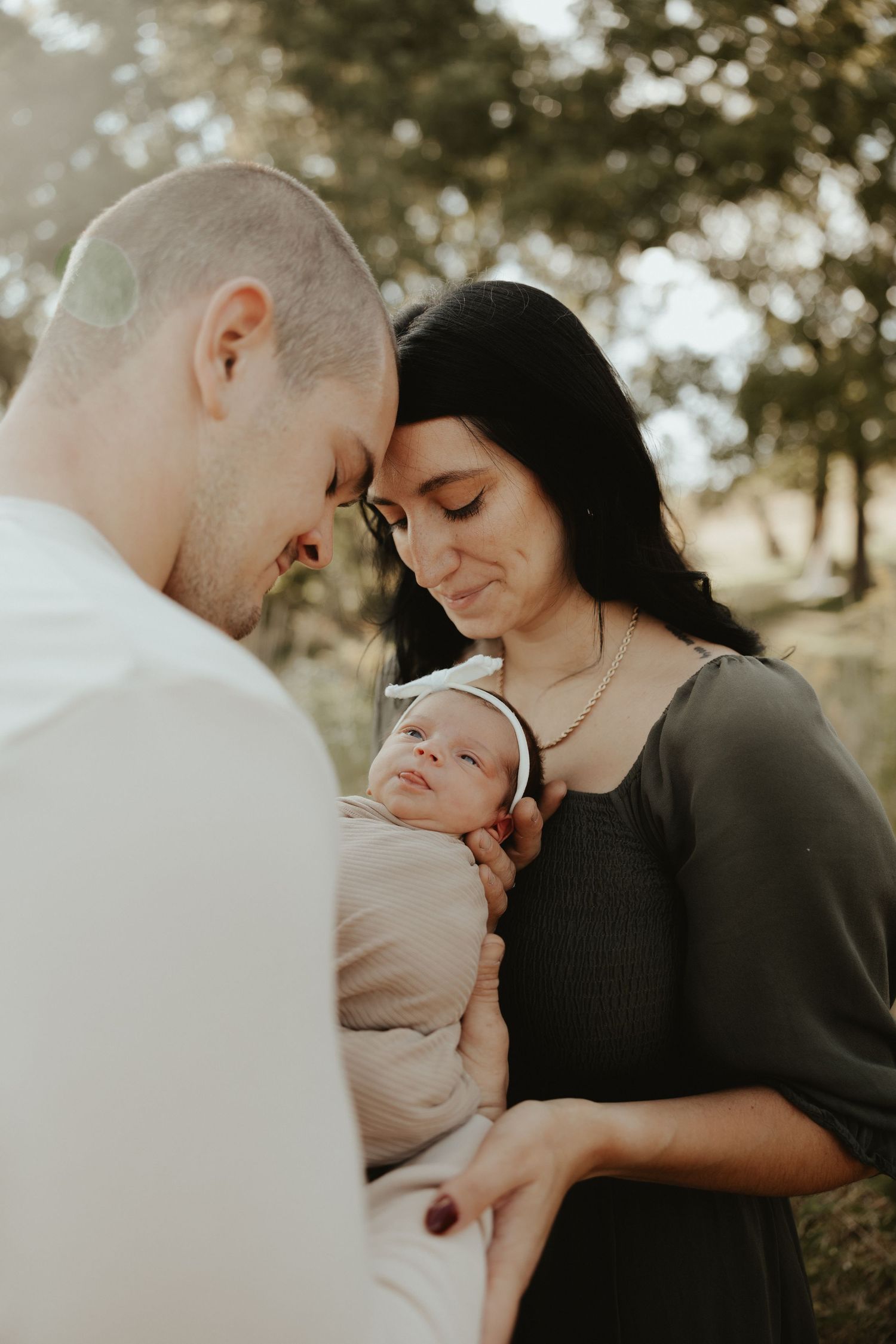 A tender family moment outdoors as parents lovingly gaze at their newborn baby in soft sunlit lighting.