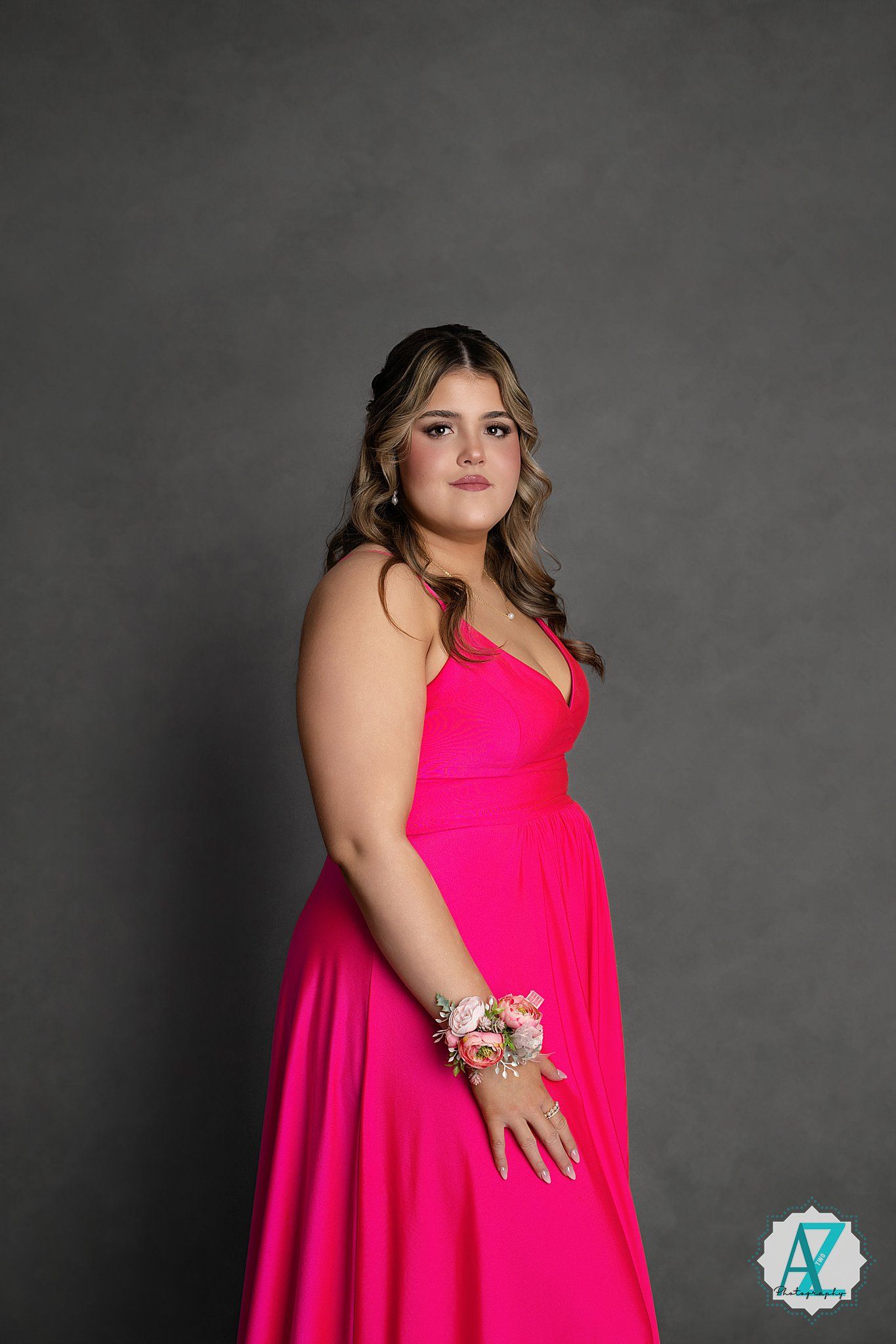 A person in a bright pink formal gown poses against a dark gray backdrop wearing a floral wrist corsage.