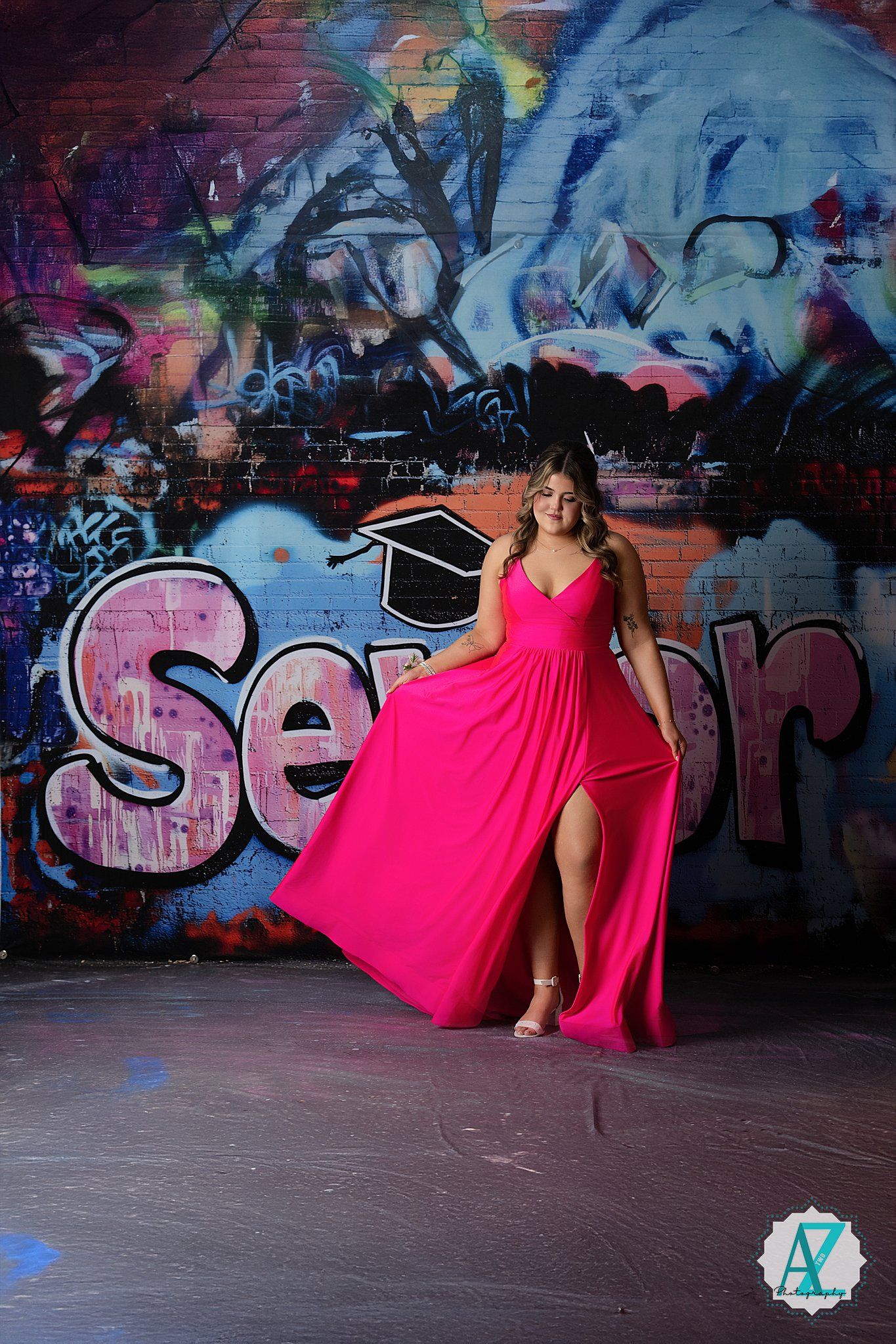 A bright pink formal gown flows dramatically against a vibrant graffiti wall backdrop featuring blue and multicolored artwork.