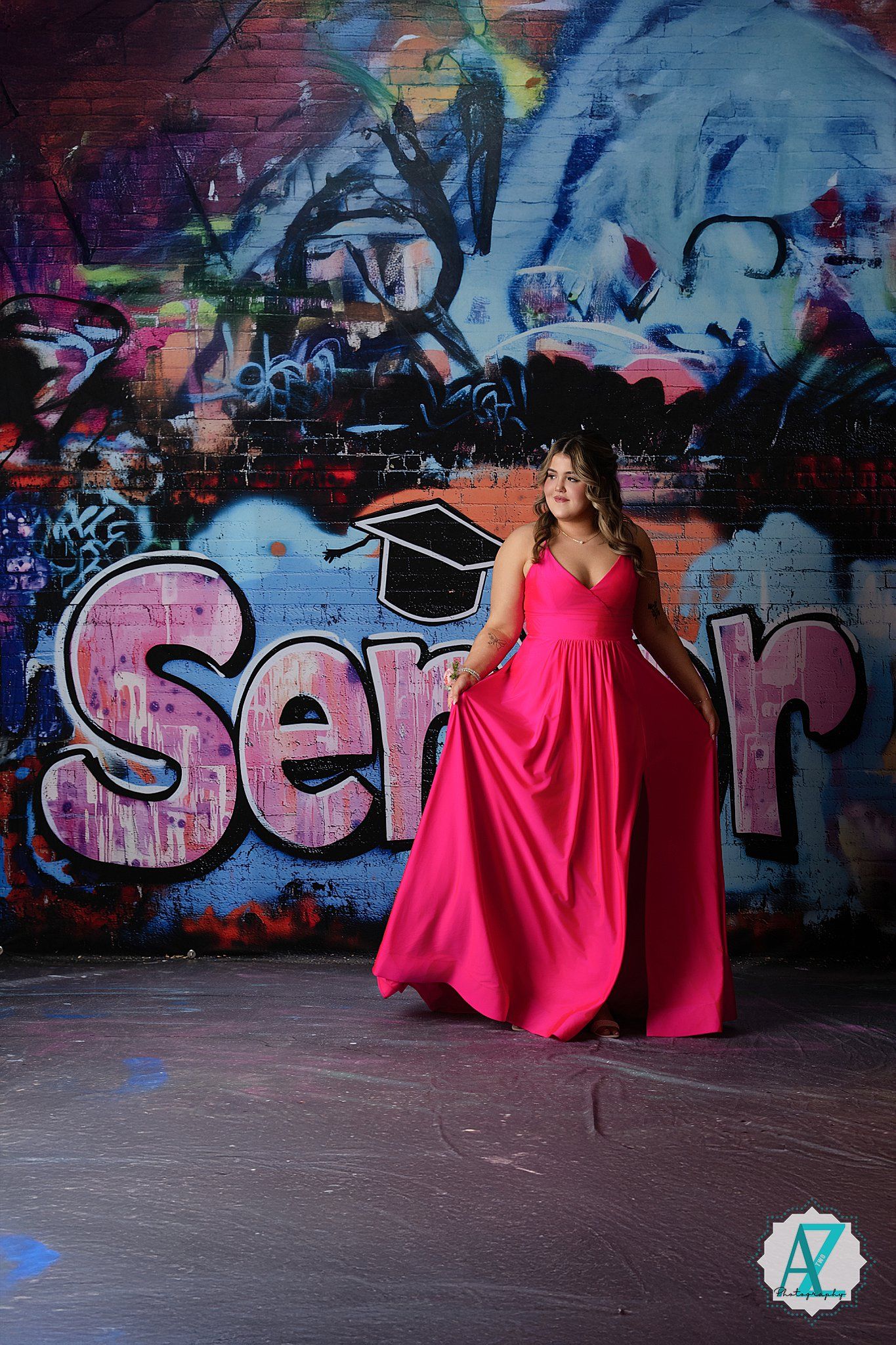 A graduate posing in a vibrant pink flowing gown against a colorful graffiti wall featuring blue and pink street art.