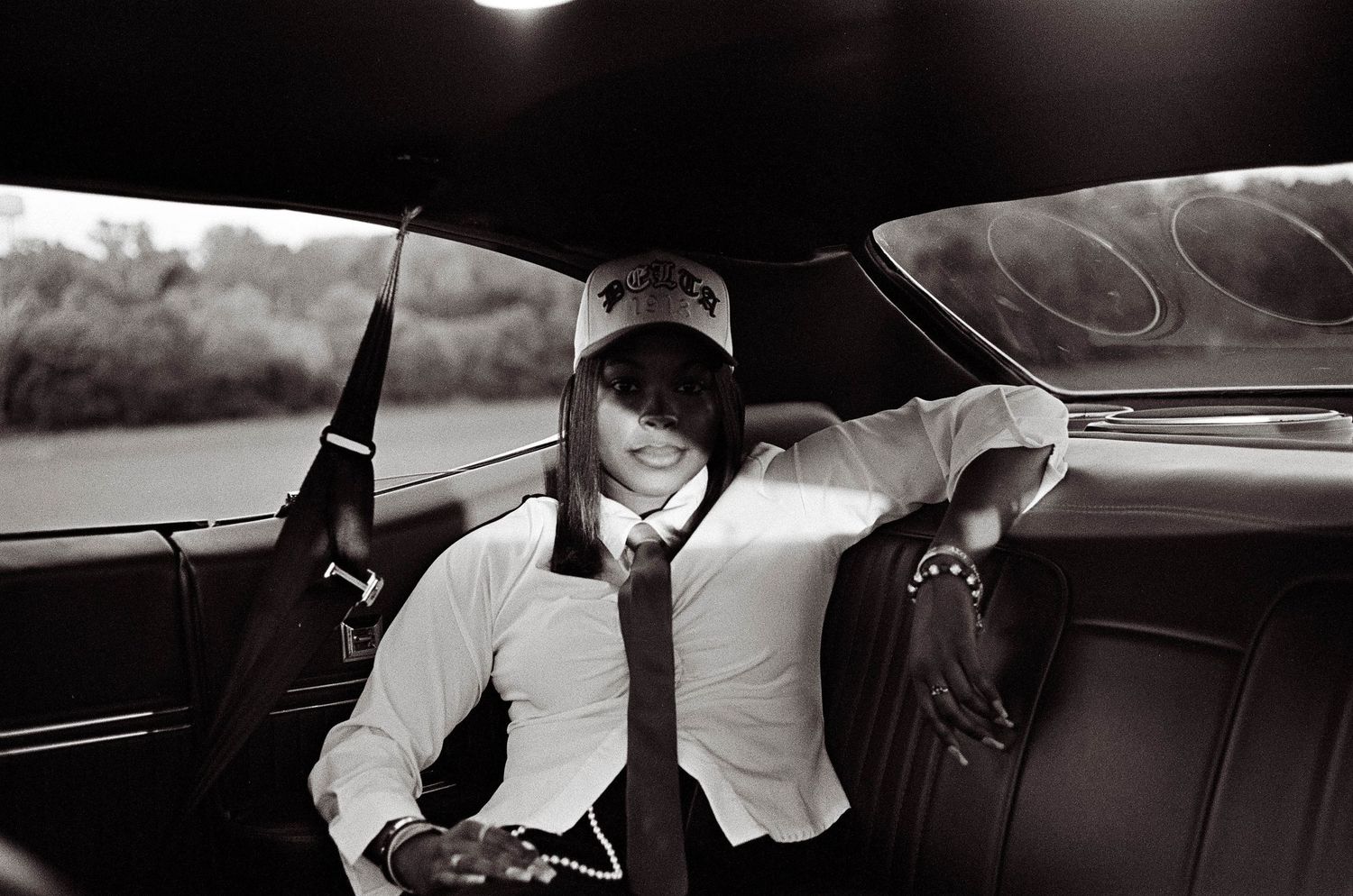 Black and white photo of person in white shirt and cap posing in vintage car interior.