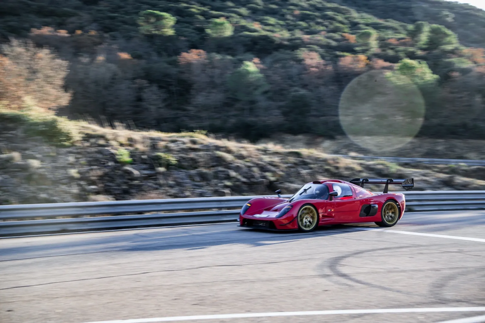 Une voiture de sport rouge roule à vive allure sur un circuit bordé de collines et de pins, dans une lumière douce de fin de journée.