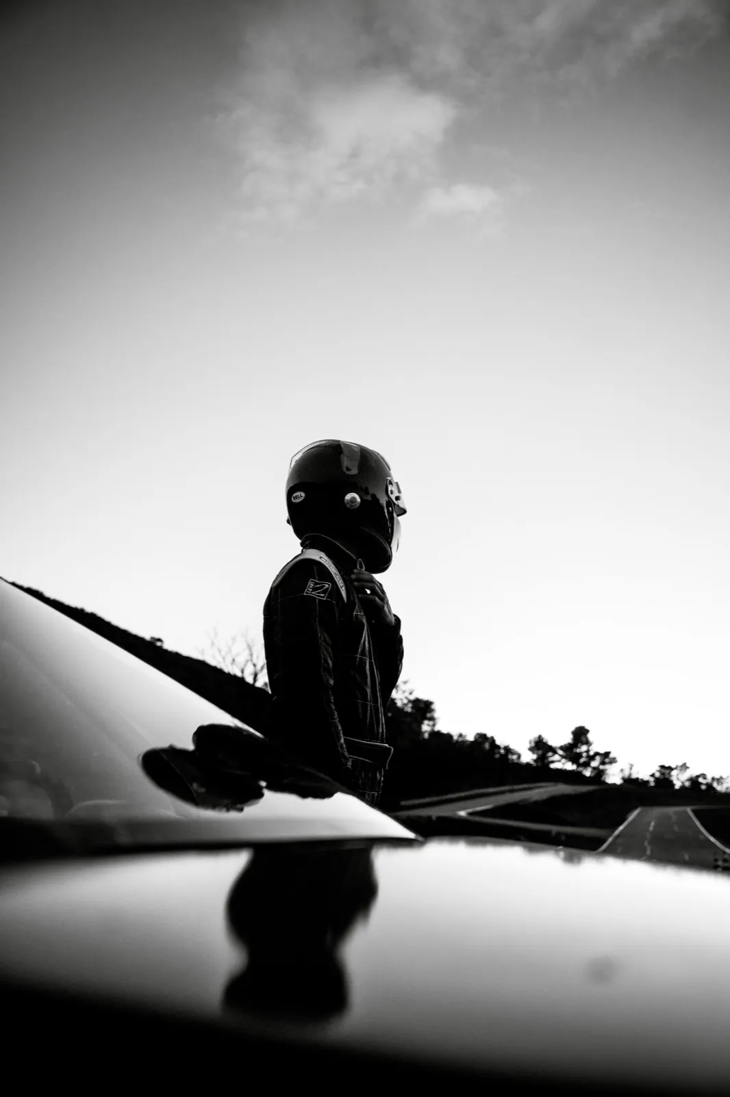 Un homme derrière le capot d’une voiture, capturé en noir et blanc avec un fort contraste lumineux.