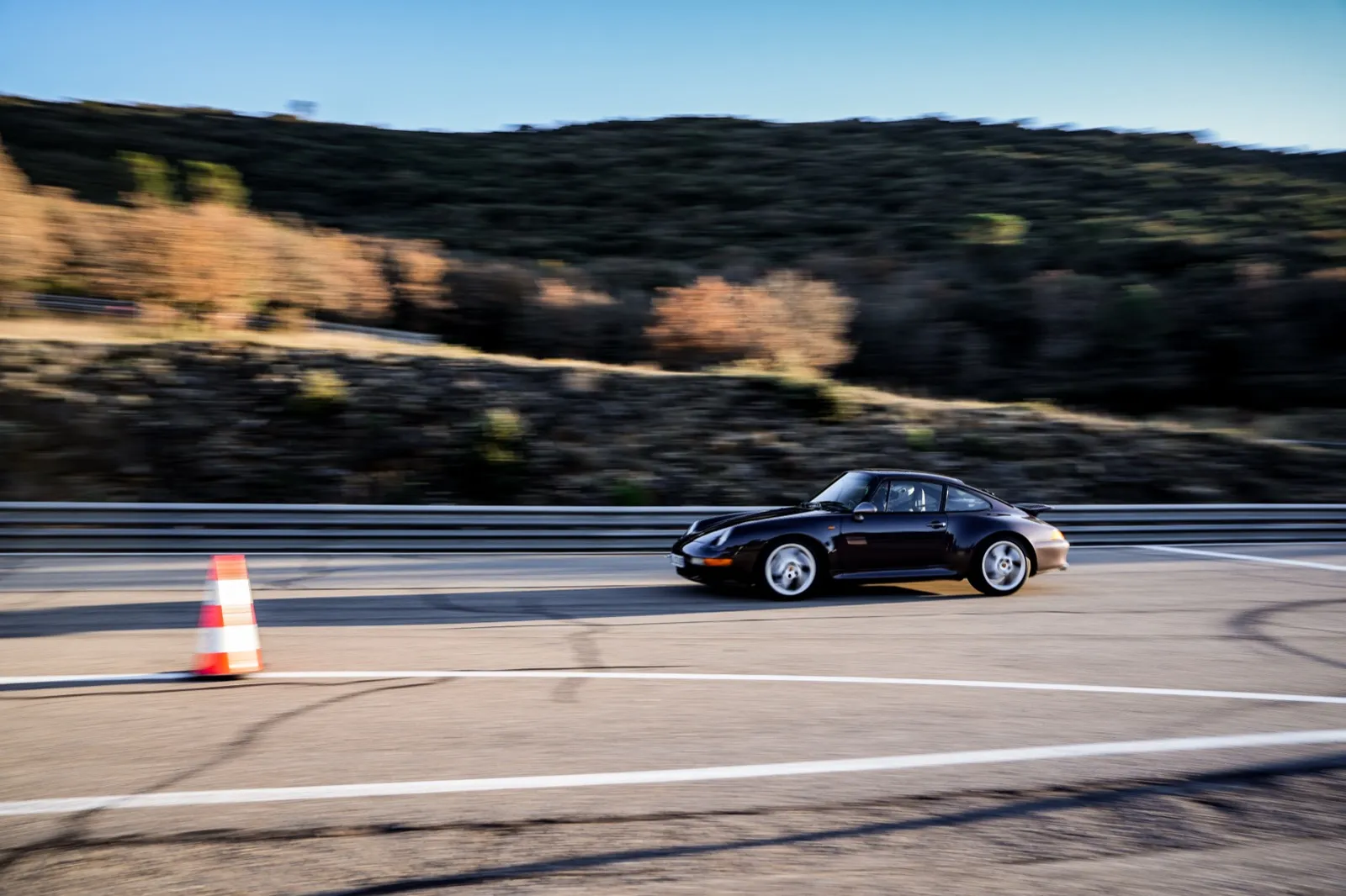 Une voiture de sport noire roule rapidement sur un circuit sinueux en pleine nature, un plot orange marquant la trajectoire.