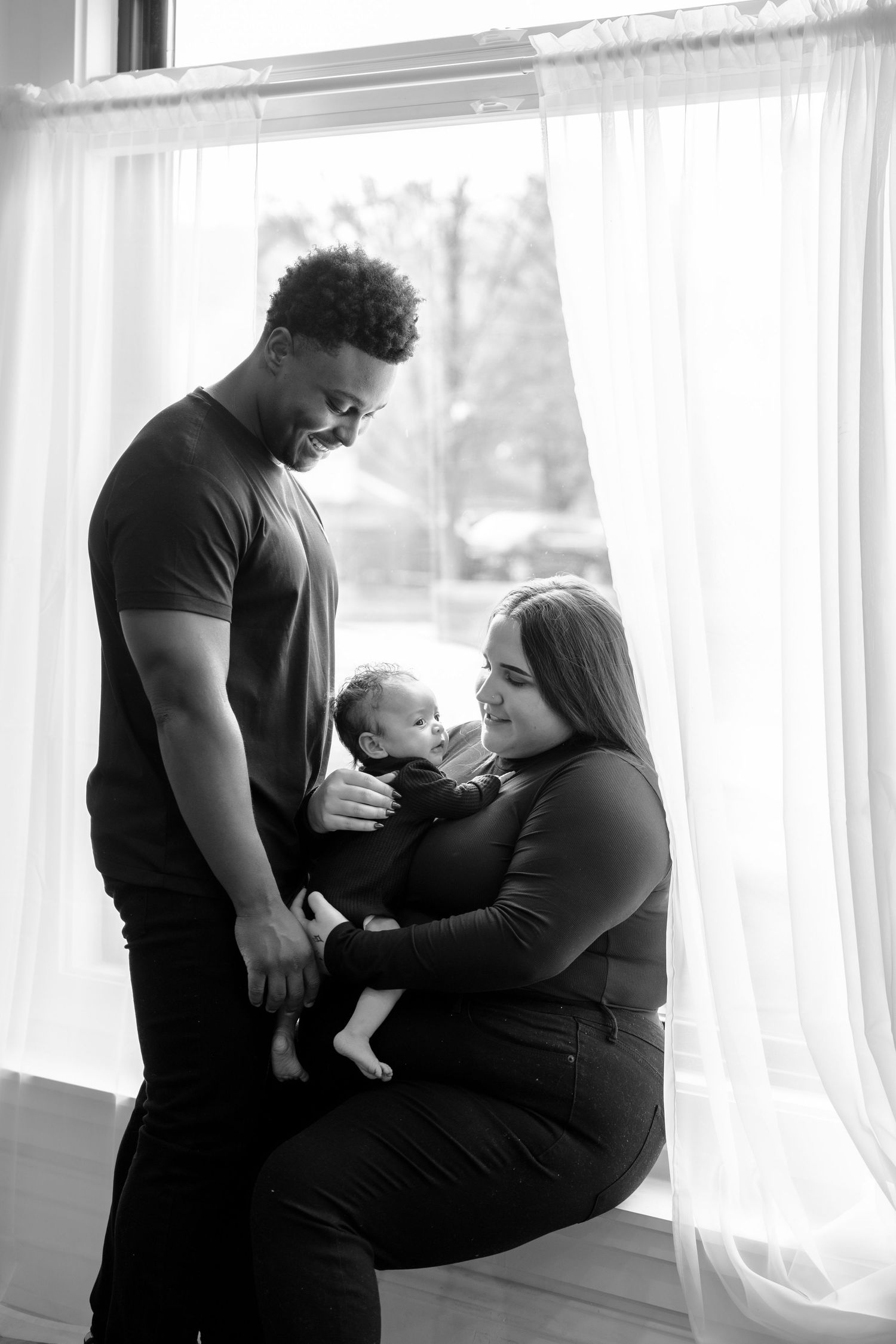 Family of three shares intimate moment by window in black and white portrait.