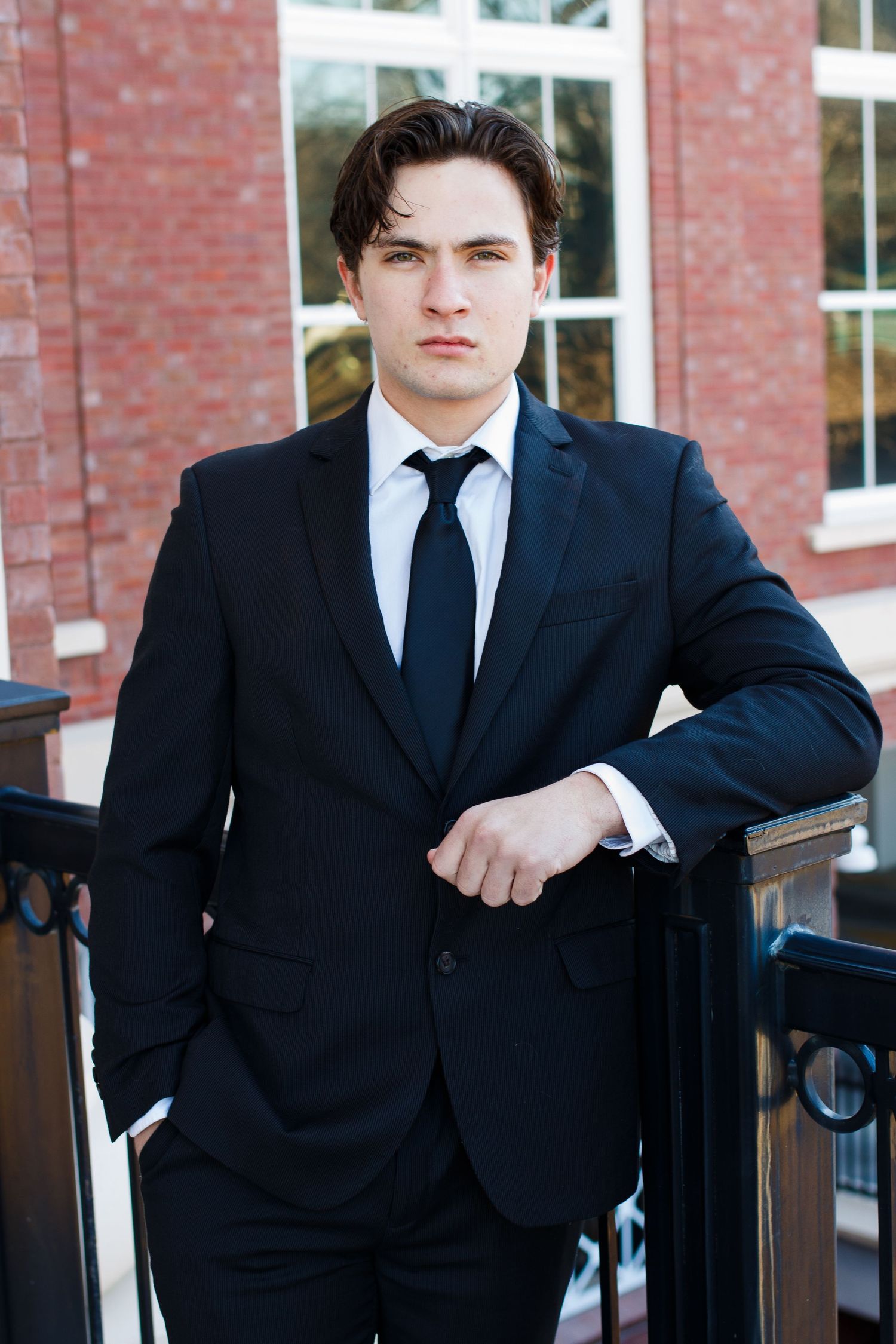 Professional portrait of a person in a black suit and tie standing by a brick building.