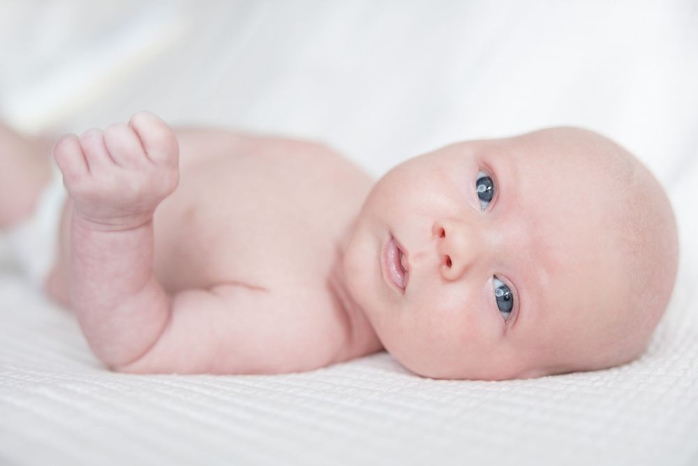 A small baby with blue eyes lying on white bedding, with one arm raised.