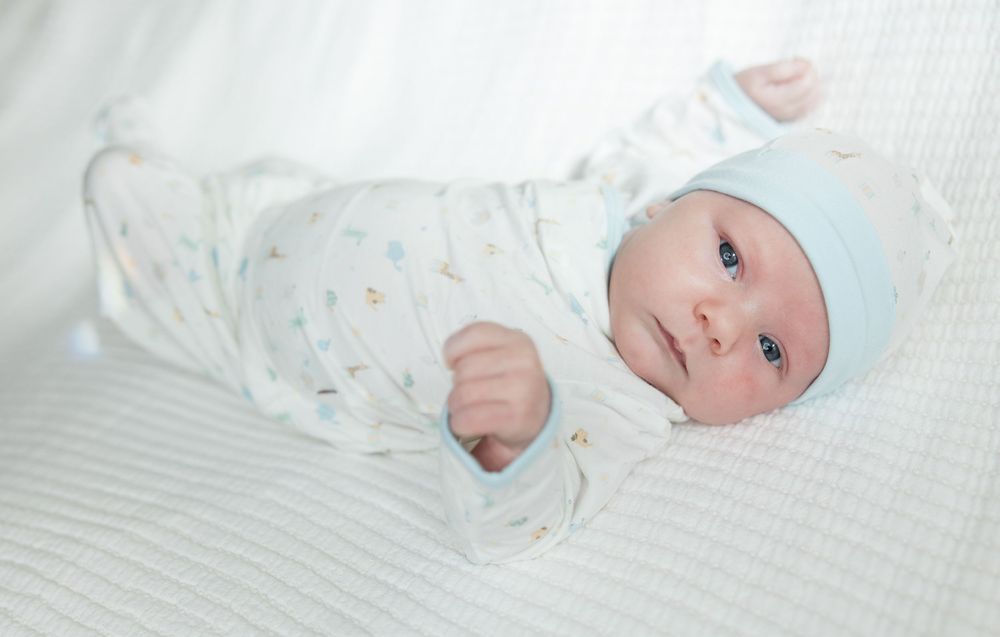Baby wearing white patterned sleeper and light blue hat rests on white textured bedding.